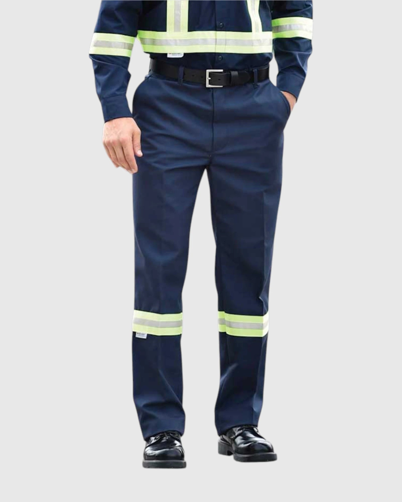 A model wearing navy blue high-visibility work pants with reflective yellow-green stripes near the knees and lower legs, paired with a matching high-visibility long-sleeve shirt and black shoes, against a light gray background.