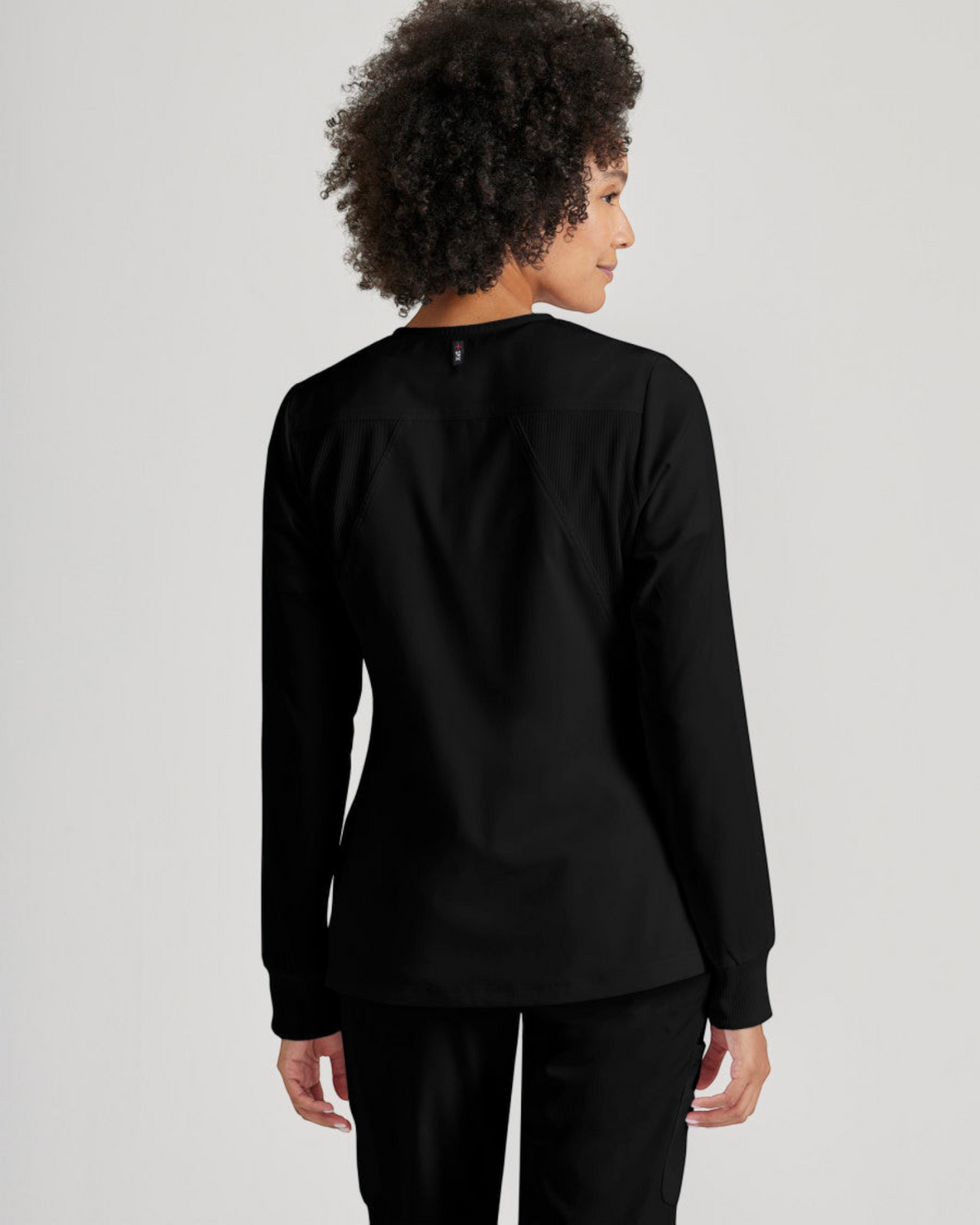 Back view of a woman wearing a black Lasalle Uniform Grey's Anatomy by Barco Gianna warm-up scrub jacket with rib-knit details.