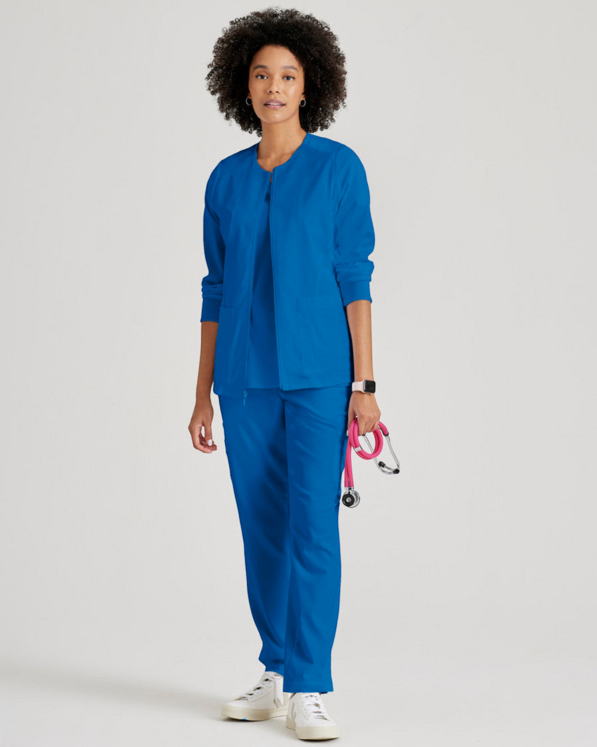 A model wearing a blue Lasalle Uniform Gianna warm-up scrub jacket with a round neck, zip front, and rib-knit cuffs.