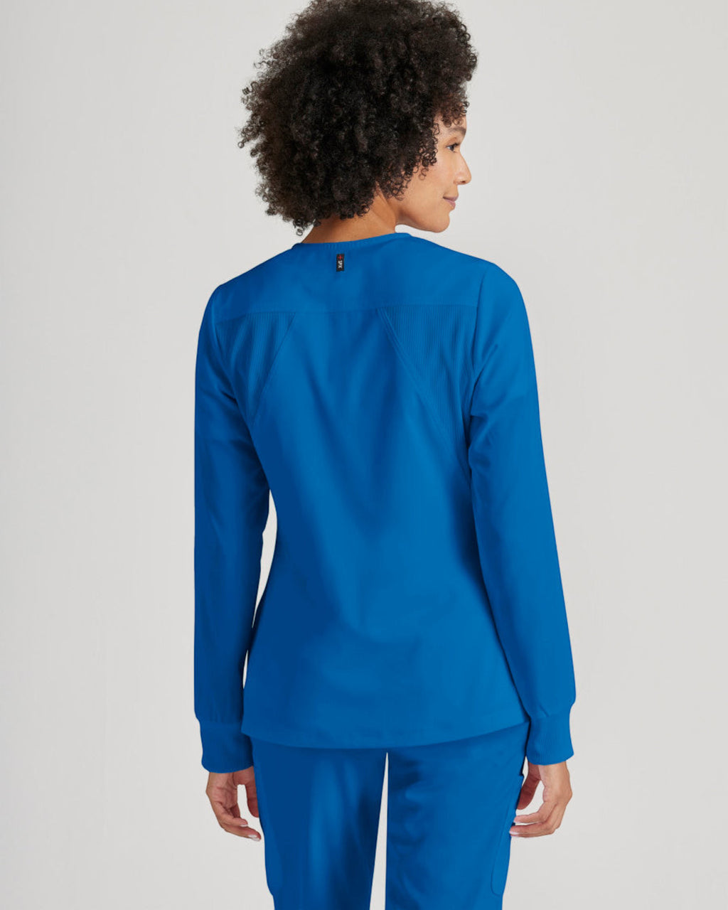 Back view of a woman wearing a royal blue Lasalle Uniform Gianna warm-up scrub jacket with rib-knit cuffs.