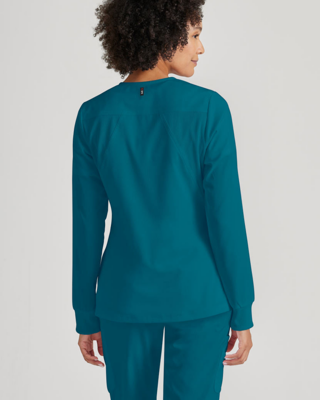 Back view of a woman wearing a teal Lasalle Uniform Gianna warm-up scrub jacket with rib-knit cuffs.