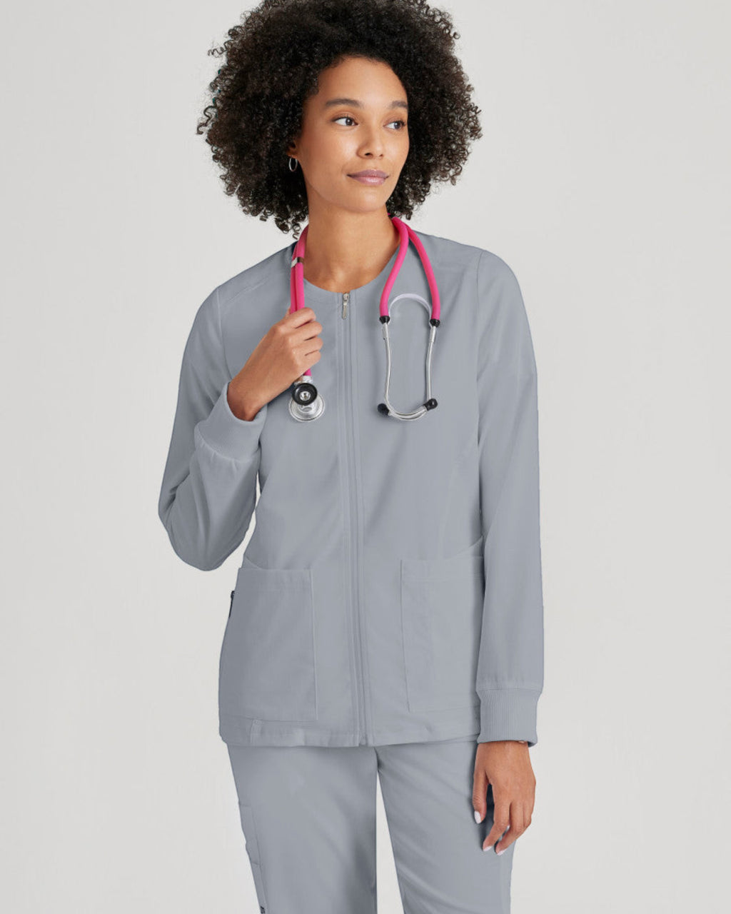 Model wearing a grey Lasalle Uniform Gianna warm-up scrub jacket with a round neck, zip front, and rib-knit cuffs.