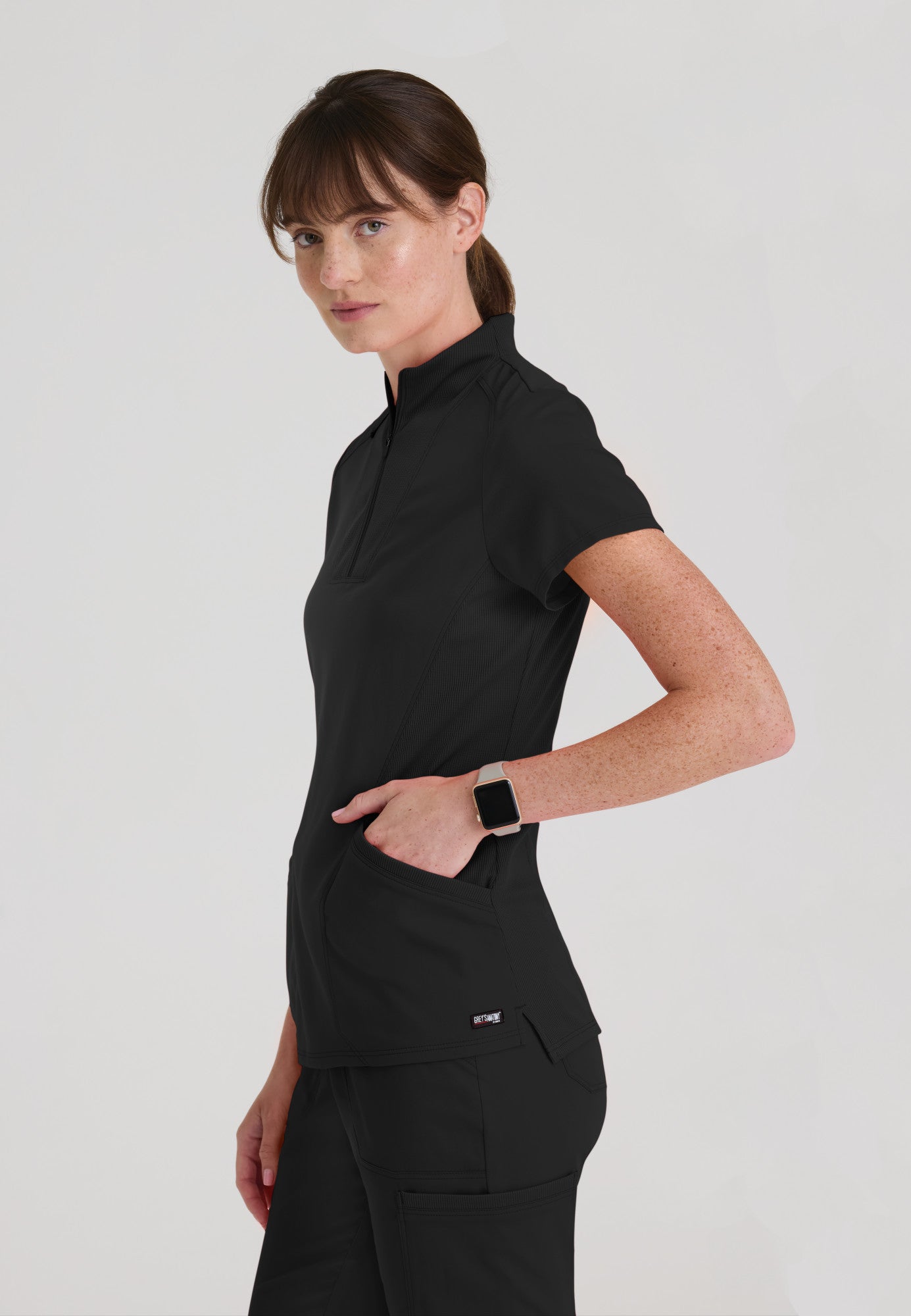 Side view of Grey's Anatomy Kristi scrub top showing flexible rib-knit stretch panels and side vents for maximum mobility. Black colour.