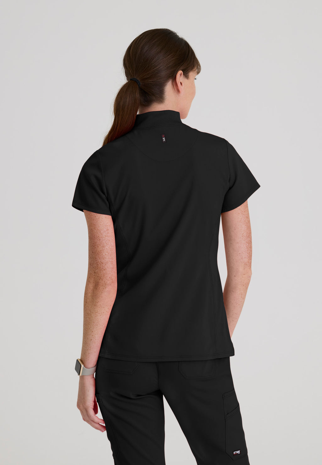Back view of Grey's Anatomy Kristi scrub top showing a high-low hemline, flexible rib-knit side panels, and tailored modern fit. Black colour.