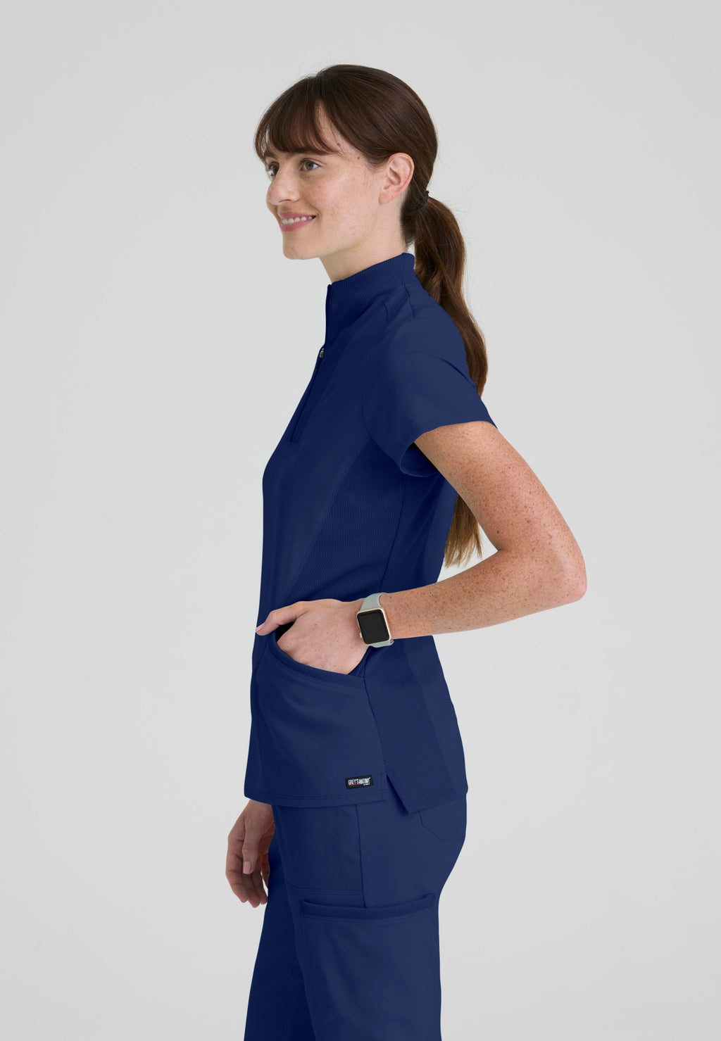 Side view of Grey's Anatomy Kristi scrub top showing flexible rib-knit stretch panels and side vents for maximum mobility. Indigo colour.