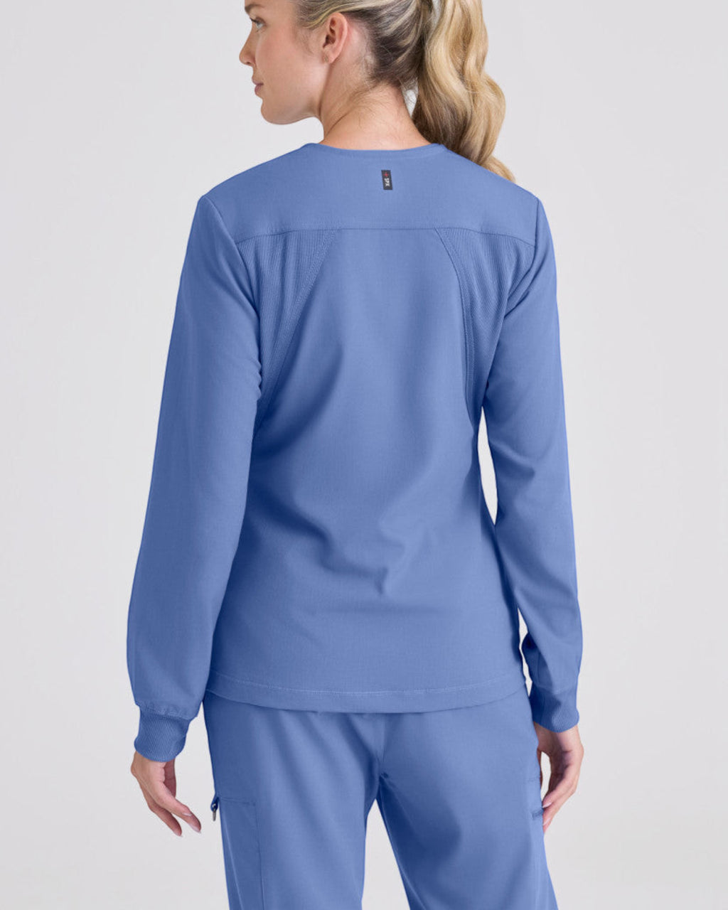 Back view of a woman wearing a blue Lasalle Uniform Gianna warm-up scrub jacket with rib-knit cuffs and stretch fabric.