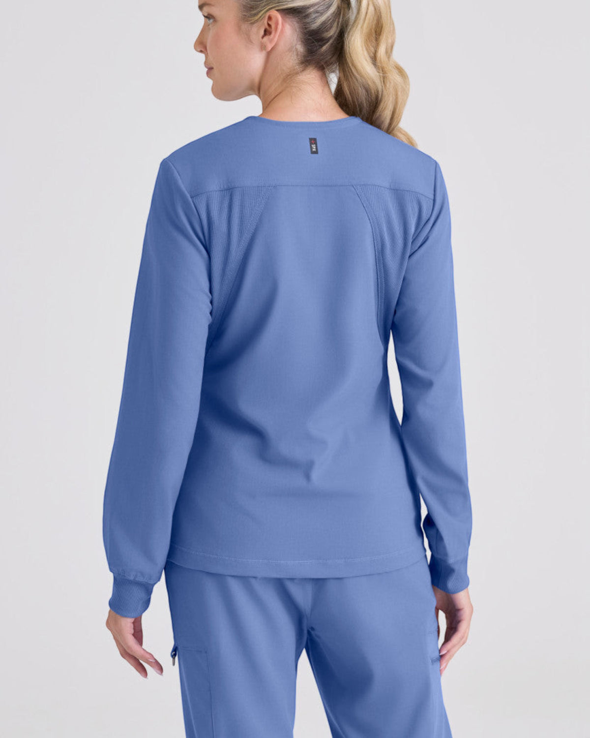 Back view of a woman wearing a blue Lasalle Uniform Gianna warm-up scrub jacket with rib-knit cuffs and stretch fabric.