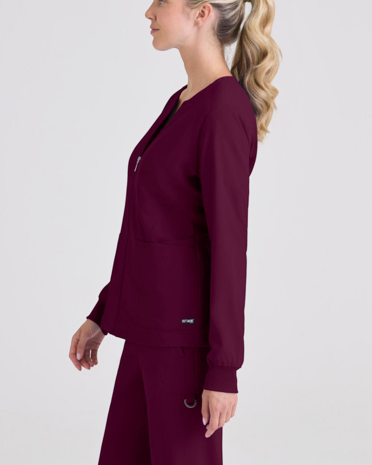 Side view of a woman wearing a burgundy Lasalle Uniform Gianna warm-up scrub jacket with rib-knit cuffs and zip front.