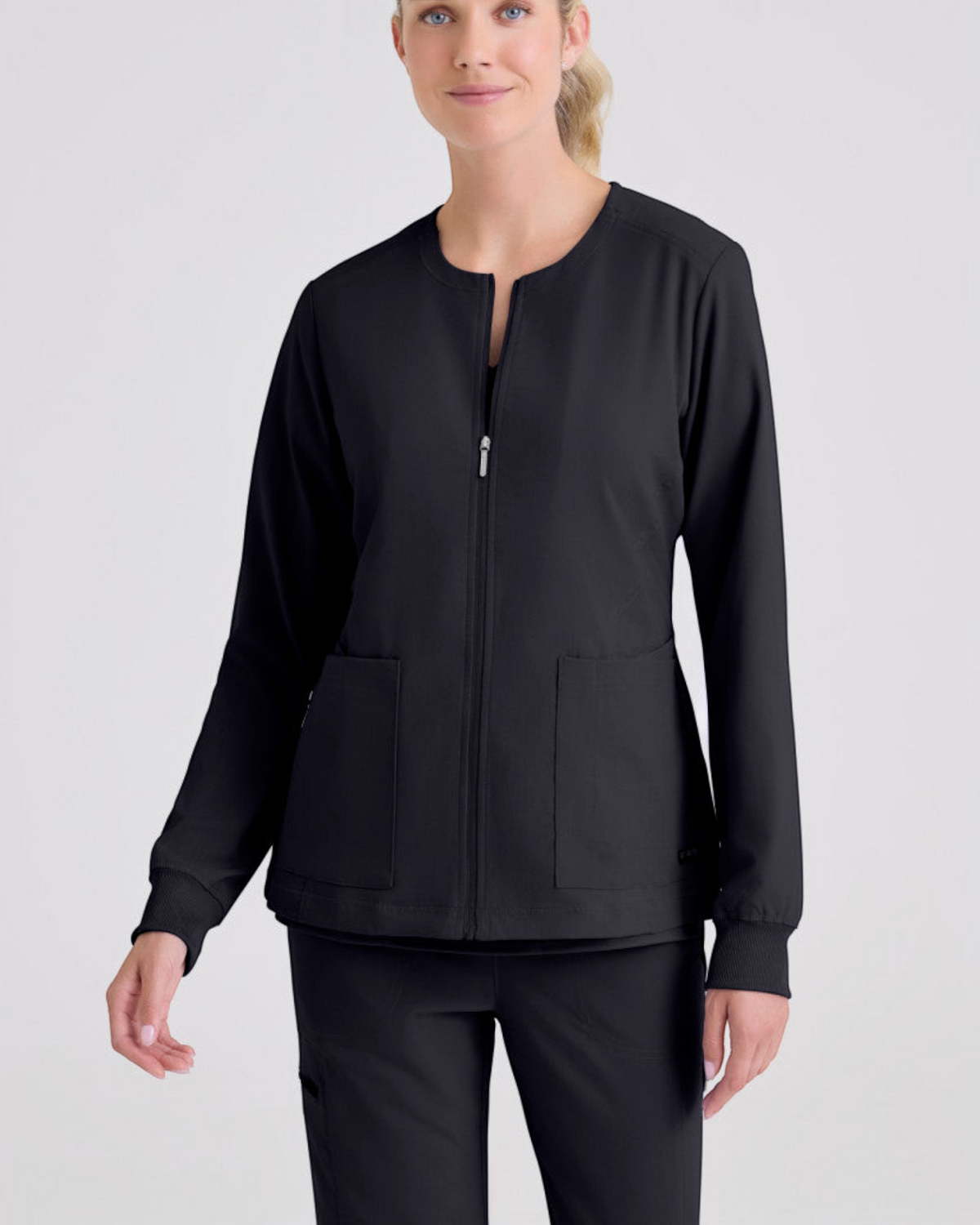 Model wearing a black Lasalle Uniform Gianna warm-up scrub jacket with a round neck, zip front, and rib-knit cuffs.
